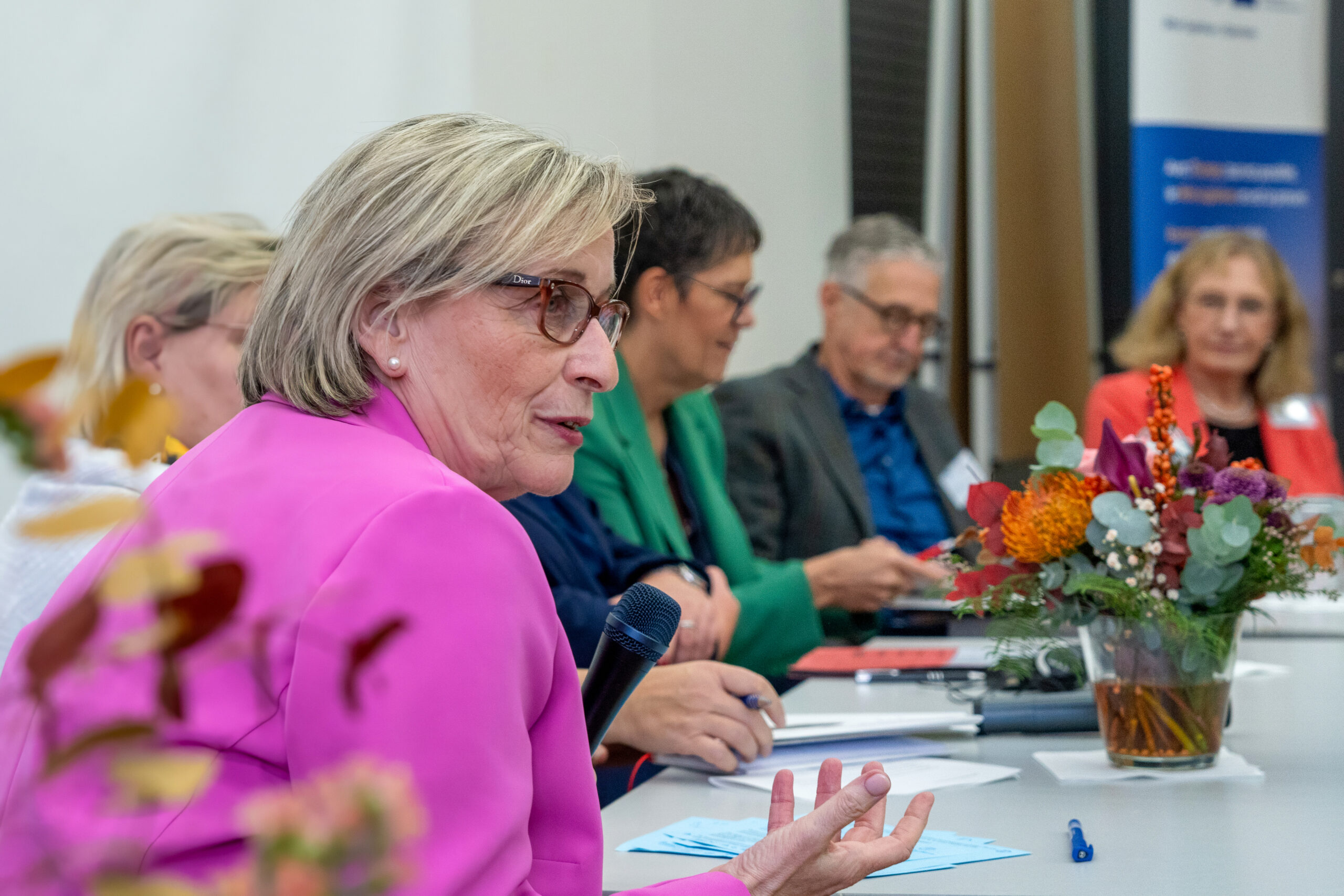 Podiumsrunde aus mehreren Personen, im Vordergrund eine Frau die in ein Mikrofon spricht.